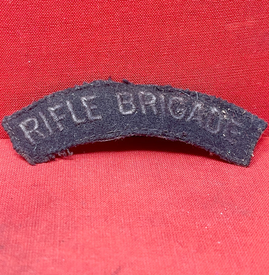 This original post-WWII Rifle Brigade shoulder title is a genuine piece of British military insignia, worn by members of the esteemed Rifle Brigade during the post-war period. Embroidered in white lettering on dark rifle green fabric, the title was typically stitched onto the upper sleeve of the battledress or service uniform. The Rifle Brigade, known for its distinguished service history and elite light infantry role, continued to uphold its proud traditions in the years following World War II. This should