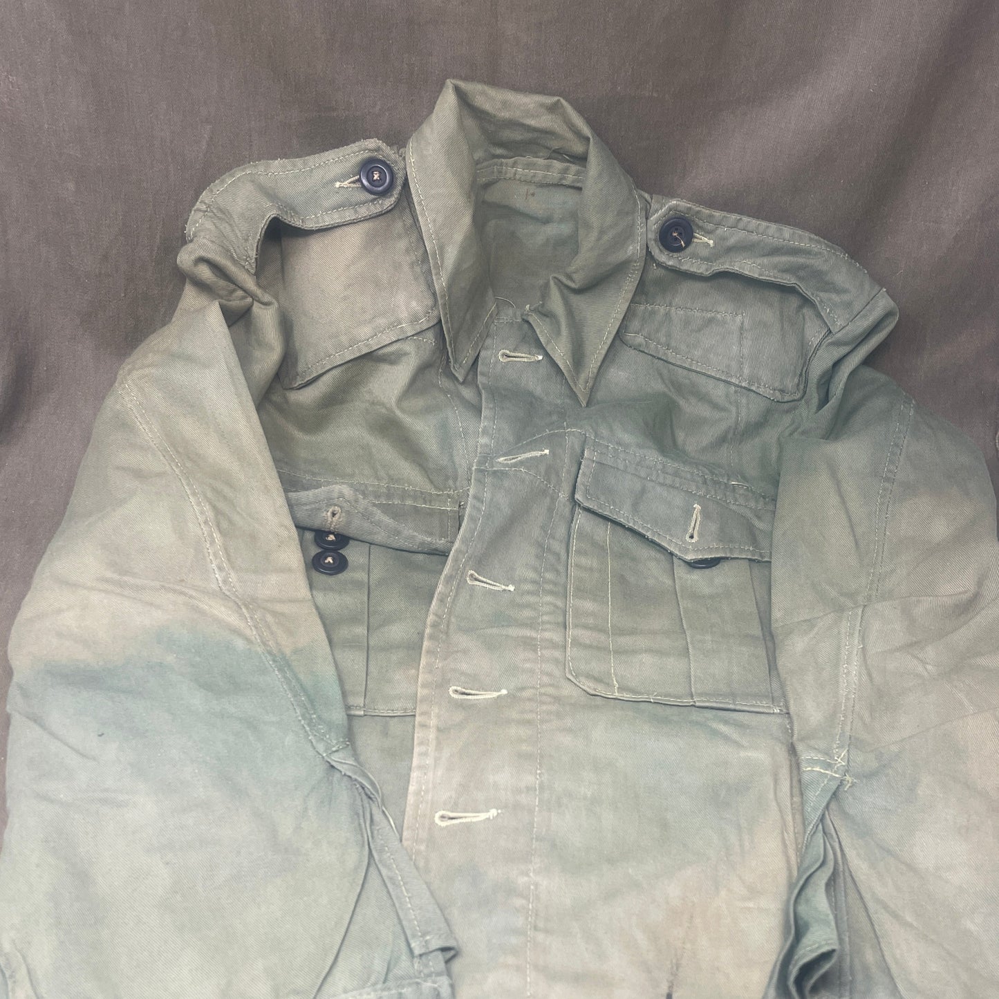 This original 1951-dated British Army Jungle Green Bush Jacket is a superb example of early post-war tropical combat clothing, manufactured by the reputable N. Lotery & Co. Ltd. Made from sturdy, breathable cotton in classic Jungle Green, this jacket was designed for use in humid and challenging environments such as Malaya and other post-war theatres of operation.
The jacket is in excellent condition, showing minimal signs of wear, with its color, stitching, and structural integrity all remarkably well-pre