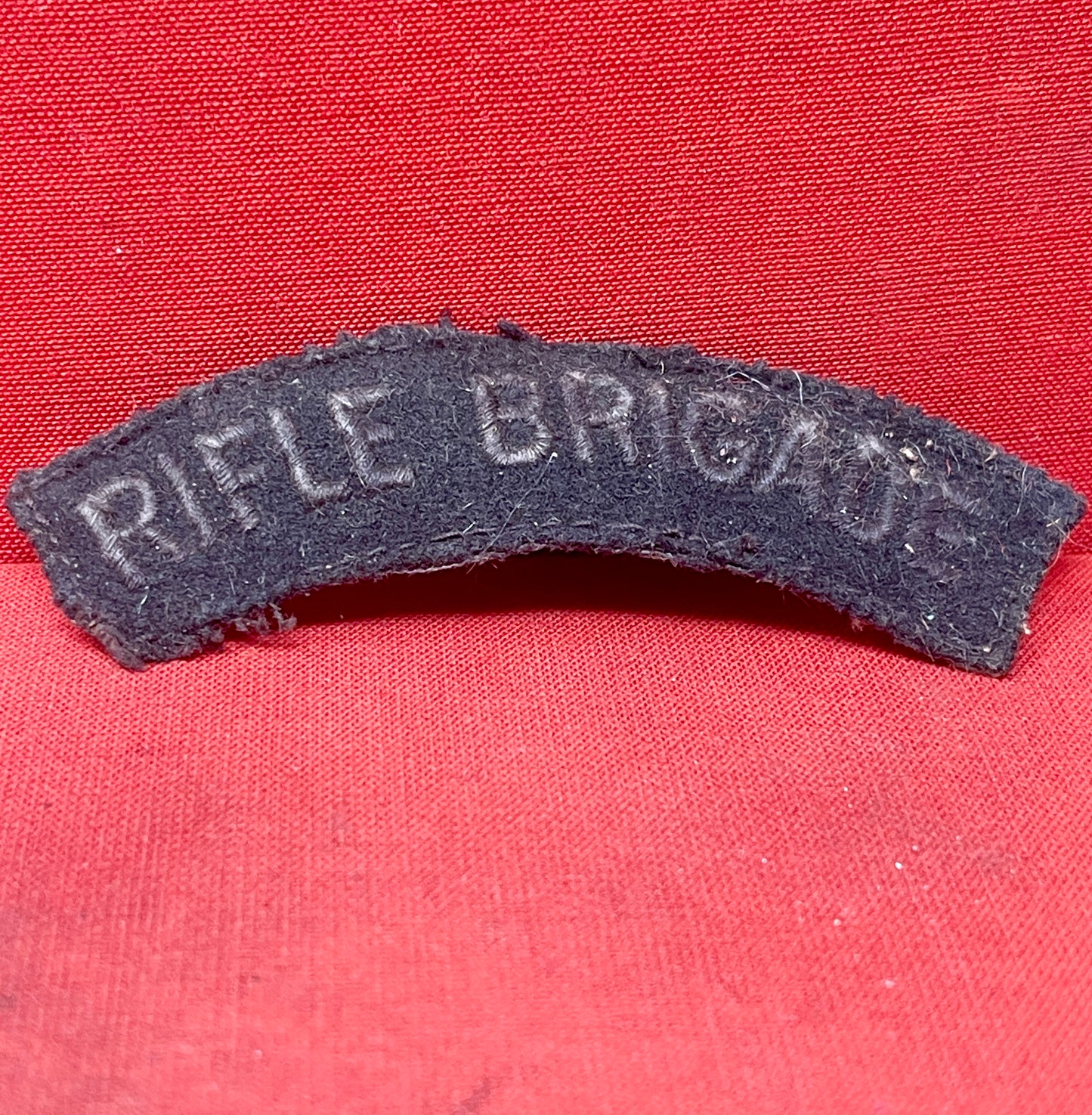 This original post-WWII Rifle Brigade shoulder title is a genuine piece of British military insignia, worn by members of the esteemed Rifle Brigade during the post-war period. Embroidered in white lettering on dark rifle green fabric, the title was typically stitched onto the upper sleeve of the battledress or service uniform. The Rifle Brigade, known for its distinguished service history and elite light infantry role, continued to uphold its proud traditions in the years following World War II. This should